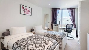 A hotel room with two beds, a desk, a chair, and a large window with curtains. - 1BD Flat with City Views, Elephant & Castle (London)