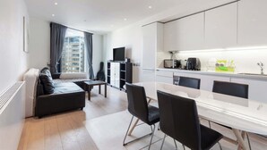 A modern hotel room with a sofa, a dining table with chairs, a kitchen area, and a large window with curtains. - 1BD Flat with City Views, Elephant & Castle (London)