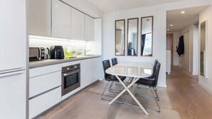 A modern kitchen with white cabinets, a built-in oven, a coffee maker, and a dining table with four chairs. - 1BD Flat with City Views, Elephant & Castle (London)