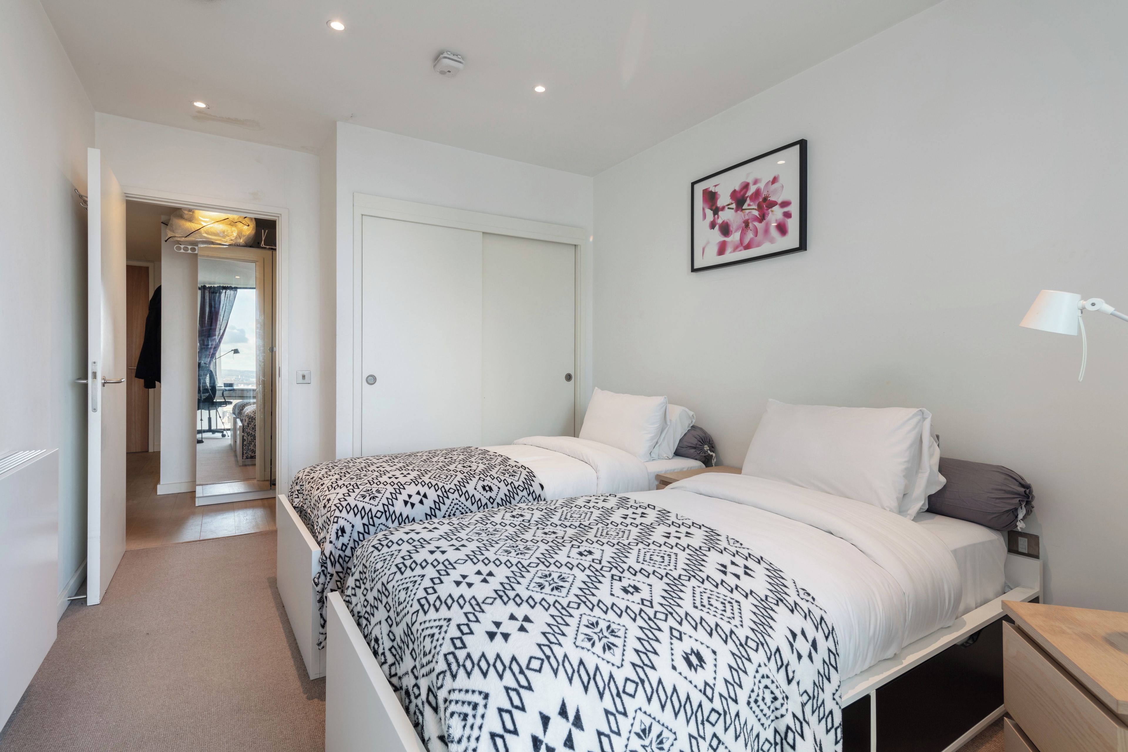 Une chambre avec deux lits, une table de chevet, une commode, un tableau encadré au mur et une porte donnant sur une autre pièce.