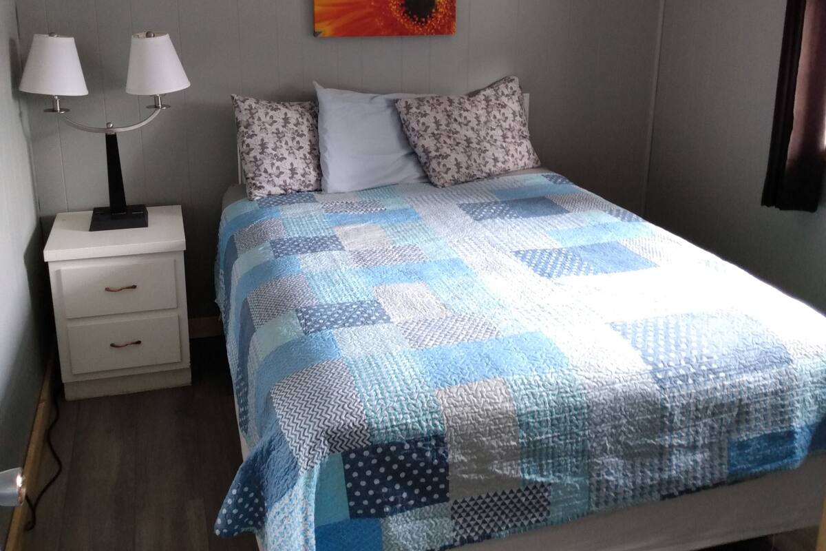 Un dormitorio con cama, mesita de noche, lámpara y un cuadro en la pared.