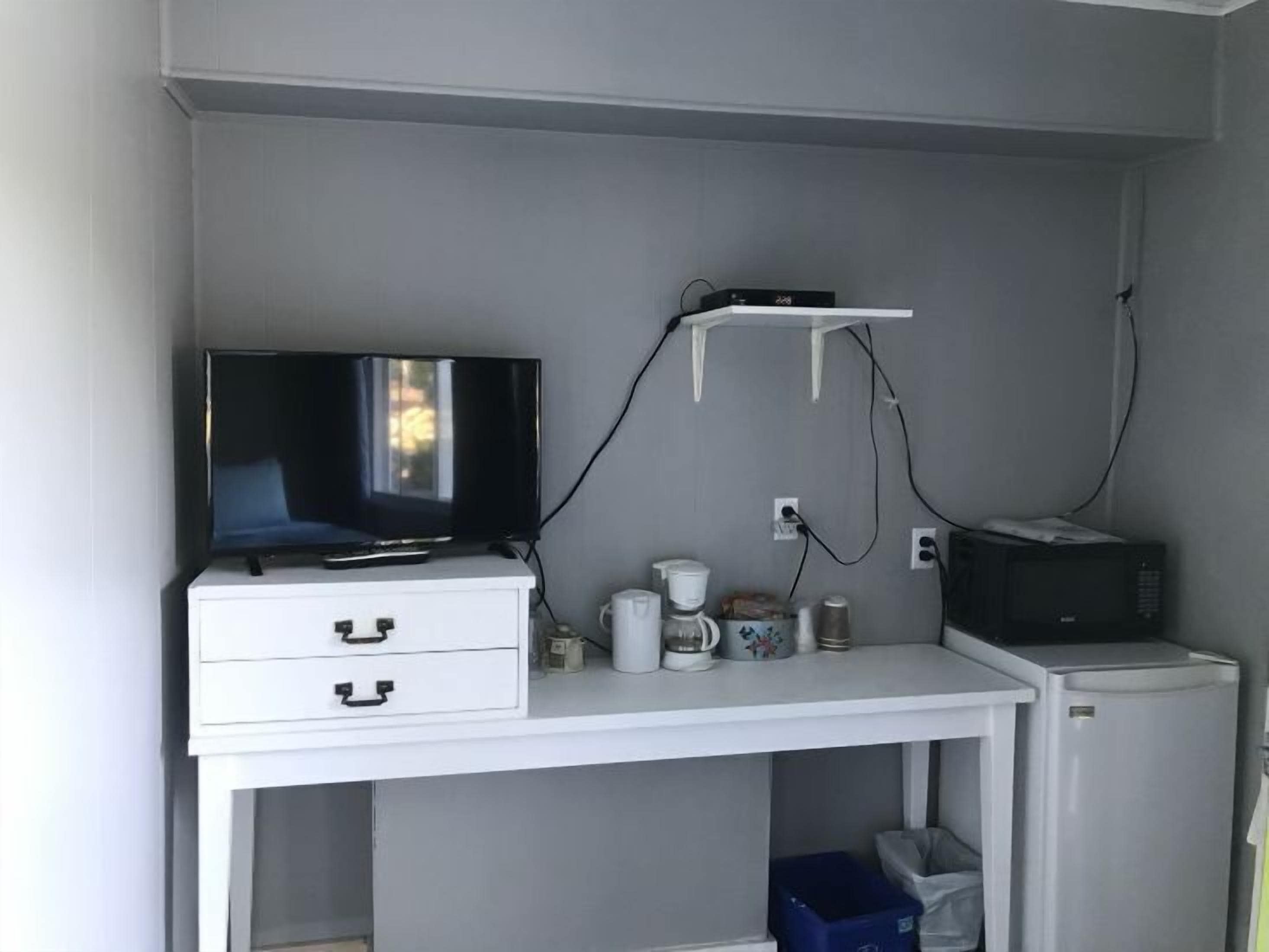 Un escritorio pequeño y blanco con televisión, cafetera, microondas y refrigerador pequeño.