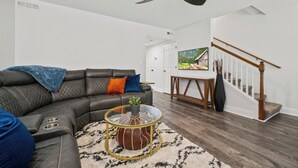 A modern living room with a sectional sofa, a glass coffee table, a ceiling fan, a staircase, and a TV in the background. - Chefs Kitchen, King Beds, Smart TVs. Close to downtown! Dead End Street (Greenville)