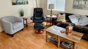 Ein Wohnzimmer mit grauem Sessel, schwarzem Ledersessel, Couchtisch aus Holz, braunem Ledersofa, einem kleinen runden Tisch mit Lampe und einer Zimmerpflanze.