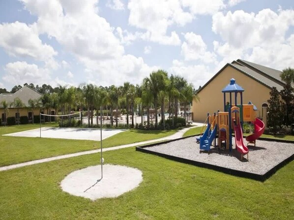 A playground with slides and a climbing structure, surrounded by a well-maintained lawn and a building with a sloped roof.