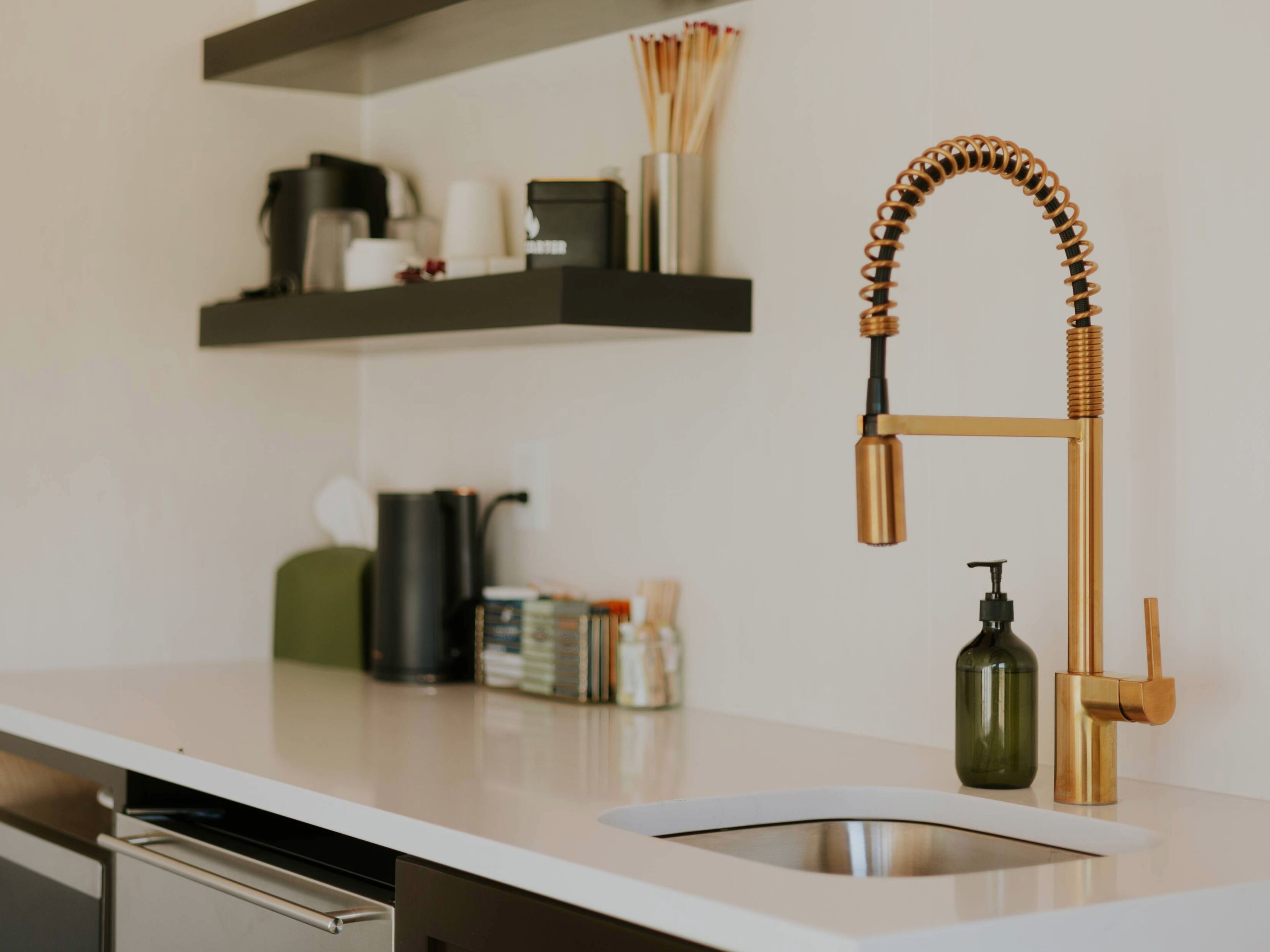 A kitchen with a white countertop, a stainless steel sink, and a gold faucet.