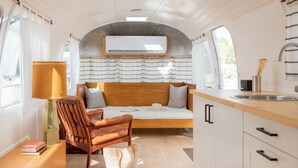 Interior compacto de una caravana con cama, una silla de madera, zona de comedor y pequeña cocina.