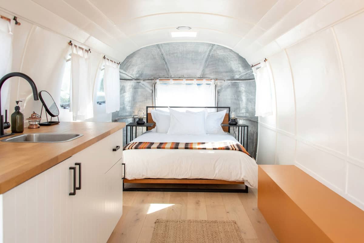 Interior compacto de uma RV com cozinha compacta, uma cama com coberta listrada e um tapete pequeno no chão.