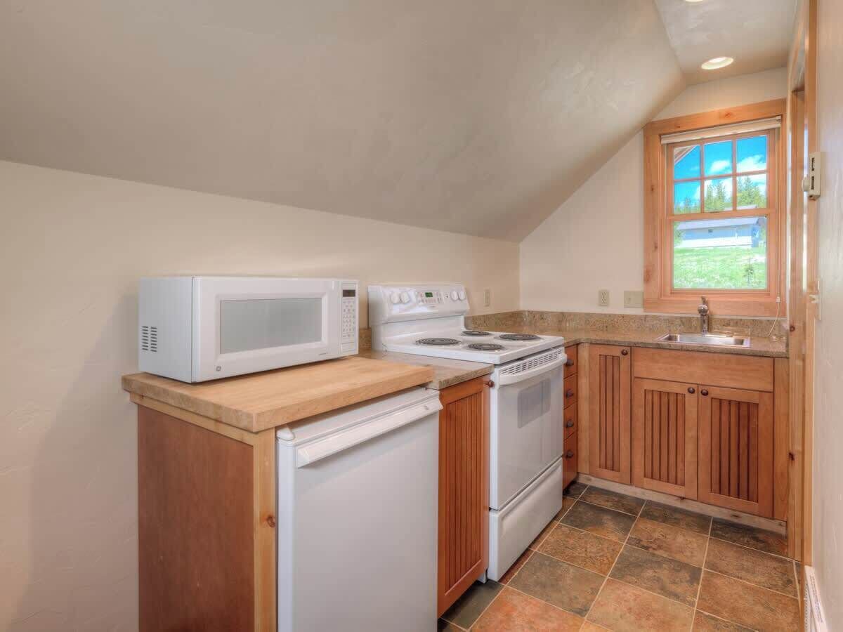 A compact kitchen with a microwave, stove, and refrigerator, wooden cabinets, and a window with a view.