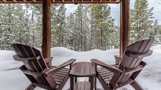 Un kiosque en bois avec deux fauteuils Adirondack et une table, entouré d’arbres couverts de neige.