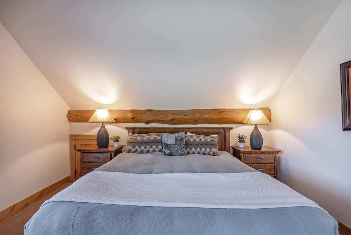 Een slaapkamer met een houten hoofdbord, twee nachtkastjes met lampen, een bed met een grijze deken en twee kussens.