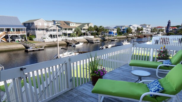 A deck with green lounge chairs and a small table, overlooking a marina with boats and houses.