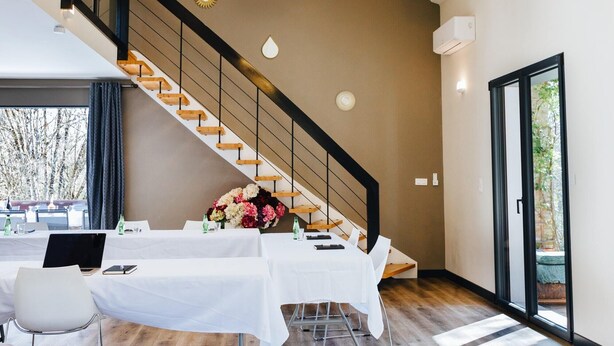 Une pièce spacieuse dotée d’un plancher en bois, d’une table blanche dressée pour le repas, d’un escalier avec une rampe noire et d’une grande fenêtre donnant sur les arbres.