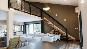Une vaste salle à manger avec une longue table dressée pour un repas, un escalier menant à un niveau supérieur et une cuisine visible au fond.