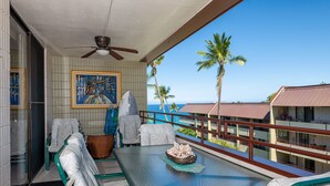 A balcony with a table, chairs, and a ceiling fan. There is a view of palm trees and a building across the water. - Stunning ✨ Comfortable and Well Decorated Property 🌺 Incredible Views! 🏖 White Sands 318 (Kailua Kona)
