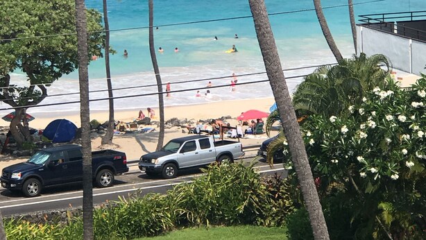 A beach view with palm trees, a road with parked vehicles, and a building with a brown roof. - Stunning ✨ Comfortable and Well Decorated Property 🌺 Incredible Views! 🏖 White Sands 318 (Kailua Kona)