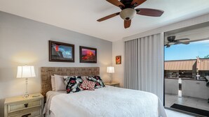 A hotel room with a large bed, two wall-mounted picture frames, a ceiling fan, and a sliding glass door to the outside. - Stunning ✨ Comfortable and Well Decorated Property 🌺 Incredible Views! 🏖 White Sands 318 (Kailua Kona)