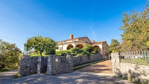 Une maison à deux étages avec une entrée surmontée d’un mur en pierre, une allée pavée et un ciel d’un bleu éclatant.