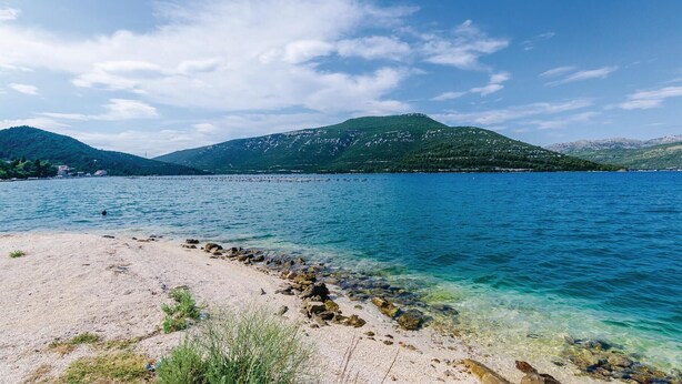 A beach with clear water, rocky shoreline, and mountains in the background.