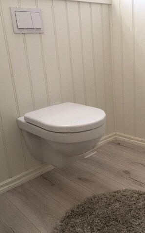 A toilet with a white lid in a room with wooden flooring and a white wall.