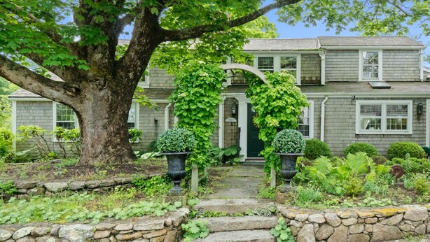 Una casa a due piani con un vialetto in pietra che conduce all'ingresso, circondata da vegetazione lussureggiante e un grande albero.