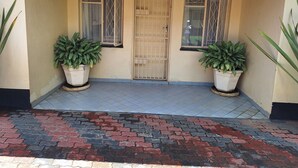 Entrada de un edificio con un porche adoquinado, dos plantas en macetas y una puerta con reja.