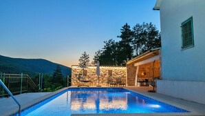 A swimming pool with a stone wall and outdoor seating area, surrounded by trees and a mountain in the background. - Awesome Home in Poljica Imotska With Wifi and 5 Bedrooms (Podbablje)