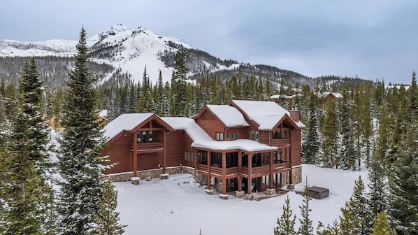 A large wooden lodge with multiple rooms, surrounded by snow-covered trees and mountains.