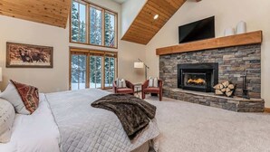 Un dormitorio con una cama grande, una chimenea de piedra, un televisor montado encima y una ventana con vistas a árboles.