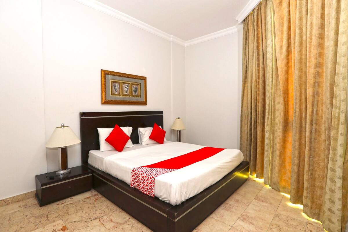 Quarto de hotel com uma cama grande, uma mesa de cabeceira com abajur, um quadro na parede e uma janela com cortinas.
