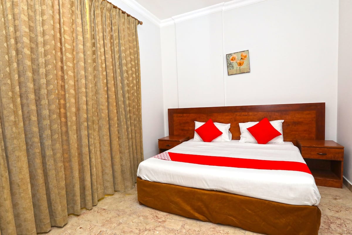 Quarto de hotel com uma cama grande, cabeceira de madeira, duas mesas de cabeceira e uma colcha vermelha e branca.