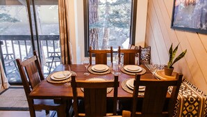 Un coin repas avec une table en bois dressée pour six personnes, une plante en pot et une vue sur les arbres par une grande fenêtre.