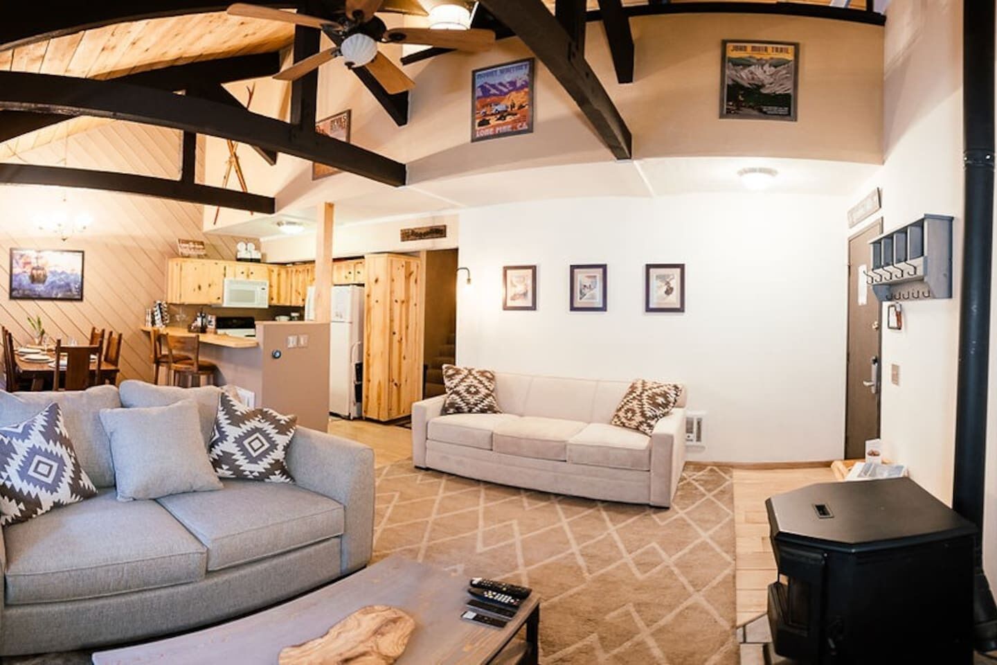 Ein gemütliches Wohnzimmer mit grauem und beigem Sofa, Holzbalken an der Decke, einem Kamin und einer Küche im Hintergrund.