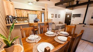 Un coin repas avec des meubles en bois, une cuisine à l’arrière-plan et un salon avec un canapé et une porte indiquant « Chambre principale ».