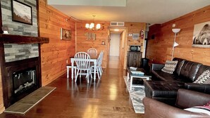 Un salón espacioso con chimenea, paredes de madera, una mesa de comedor con sillas, un sofá de cuero y un televisor.