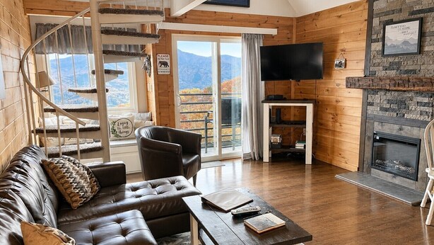 Una sala de estar acogedora con sofá, sillón y chimenea. Un televisor está montado en la pared, y hay una escalera que lleva a un balcón con vistas.