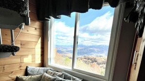 Interior acogedor de una cabaña con un banco de madera, un rascador para gatos y una ventana que ofrece una vista panorámica de montañas.