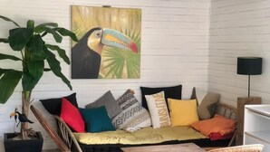 A cozy room with a wooden sofa, colorful cushions, a large potted plant, and a wall-mounted painting of a toucan. - Atypical chalet, calm nature and river (Hautecourt-Romanèche)