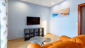 A living room with a sofa, a small round table, a TV mounted on the wall, and a painting. - Cozy Oceanview Condo w/ Swimming Pool Near the Beach (Playa Flamingo)
