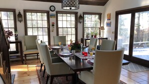 A dining area with a wooden table set for four, surrounded by chairs. There are windows with blinds, a clock on the wall, and a hanging light fixture. - Waterfront lake house  private beach dock (harwich)
