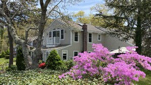A two-story house with a balcony, surrounded by lush greenery and flowering bushes. - Waterfront lake house  private beach dock (harwich)