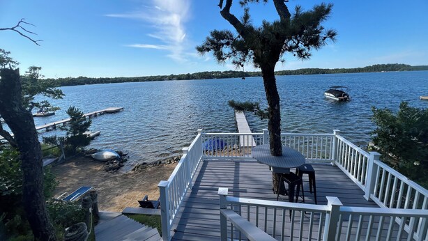 A wooden deck with a railing overlooking a lake, a dock, and a boat. - Waterfront lake house  private beach dock (harwich)