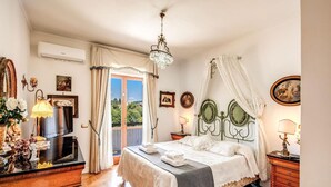 Ein Schlafzimmer mit einem Bett, einer Kandelaberleuchte, einem Schminktisch und Blick auf Grün.