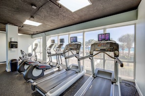A gym with treadmills, exercise bikes, and a rowing machine, with large windows offering a view of the outdoors.