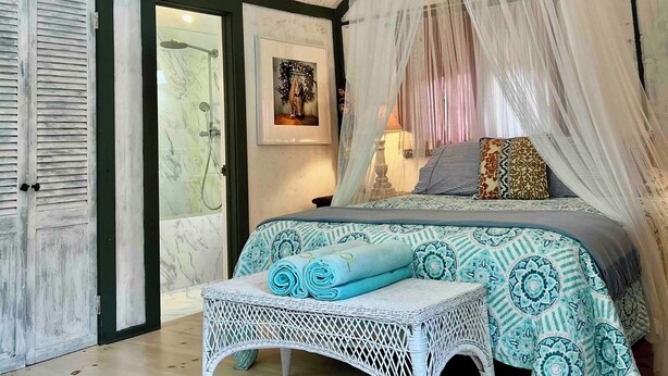 Une chambre avec un lit, un banc en osier blanc, une douche et un plafond avec des poutres apparentes.