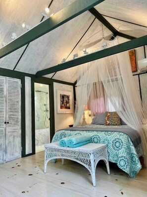 Una camera da letto con un letto, una panca in vimini bianco, una doccia e un soffitto a vista con travi.