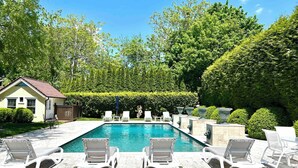Une piscine rectangulaire, bordée de chaises longues blanches disposées tout autour, et entourée de haies et d’arbres bien entretenus.