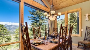 Un coin repas avec une table en bois dressée pour six personnes, entourée de chaises en bois, d’un lustre et offrant une vue sur les montagnes à travers de grandes fenêtres.