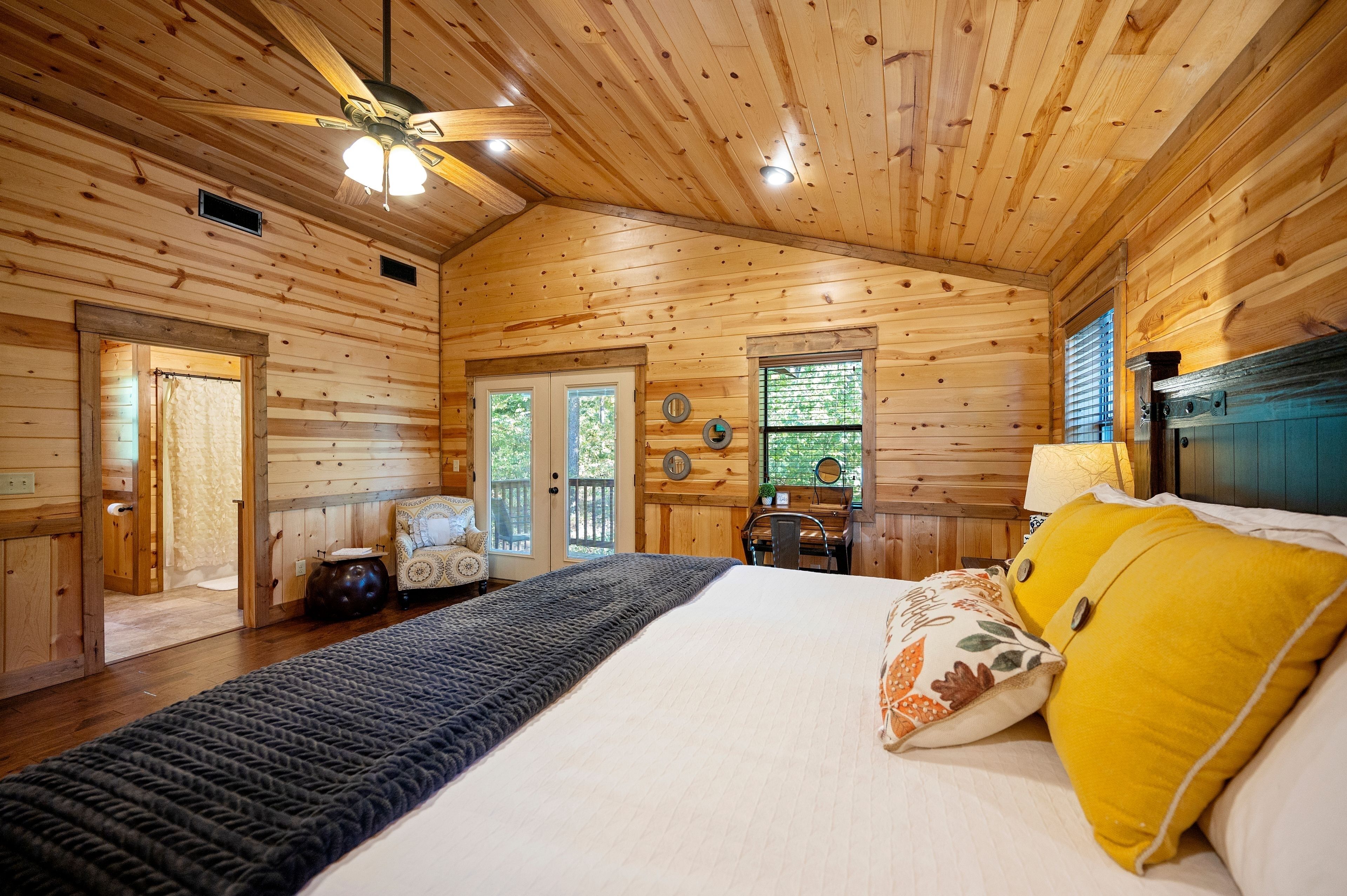 Une chambre à coucher aménagée dans un chalet en rondins, avec un lit, un ventilateur de plafond et une vue sur l’extérieur par une porte.