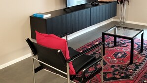 Una sala de estar moderna con un sofá negro, una mesa de centro de vidrio, un televisor de pantalla plana, un mueble de madera oscura con libros y un tapete rojo estampado.